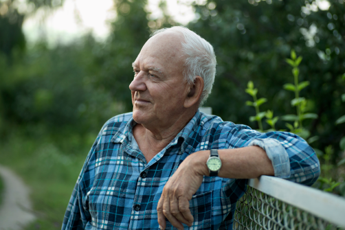 A thoughtful older adult man in a blue plaid shirt resting on a fence in a peaceful outdoor setting